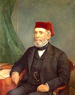 White man with grey beard, wearing suit and red fez, seated in a wooden armchair and resting his elbow near books on a table.